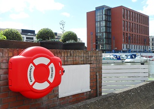 Guardian™ Lifebuoy Cabinet in red with life ring inside and white panel graphics with emergency text, situated outside hospital with marina