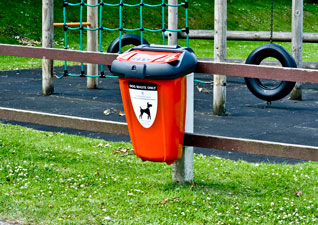 Retriever™ 50 Dog Waste Bin in red with ‘dog waste only’ sticker, fixed onto wooden fence in front of children’s playground